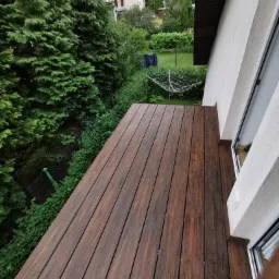 Nowy, drewniany taras z widokiem na zieleń ogrodu, z suszarką na pranie w tle, przylegający do białego budynku z widocznym fragmentem okna.