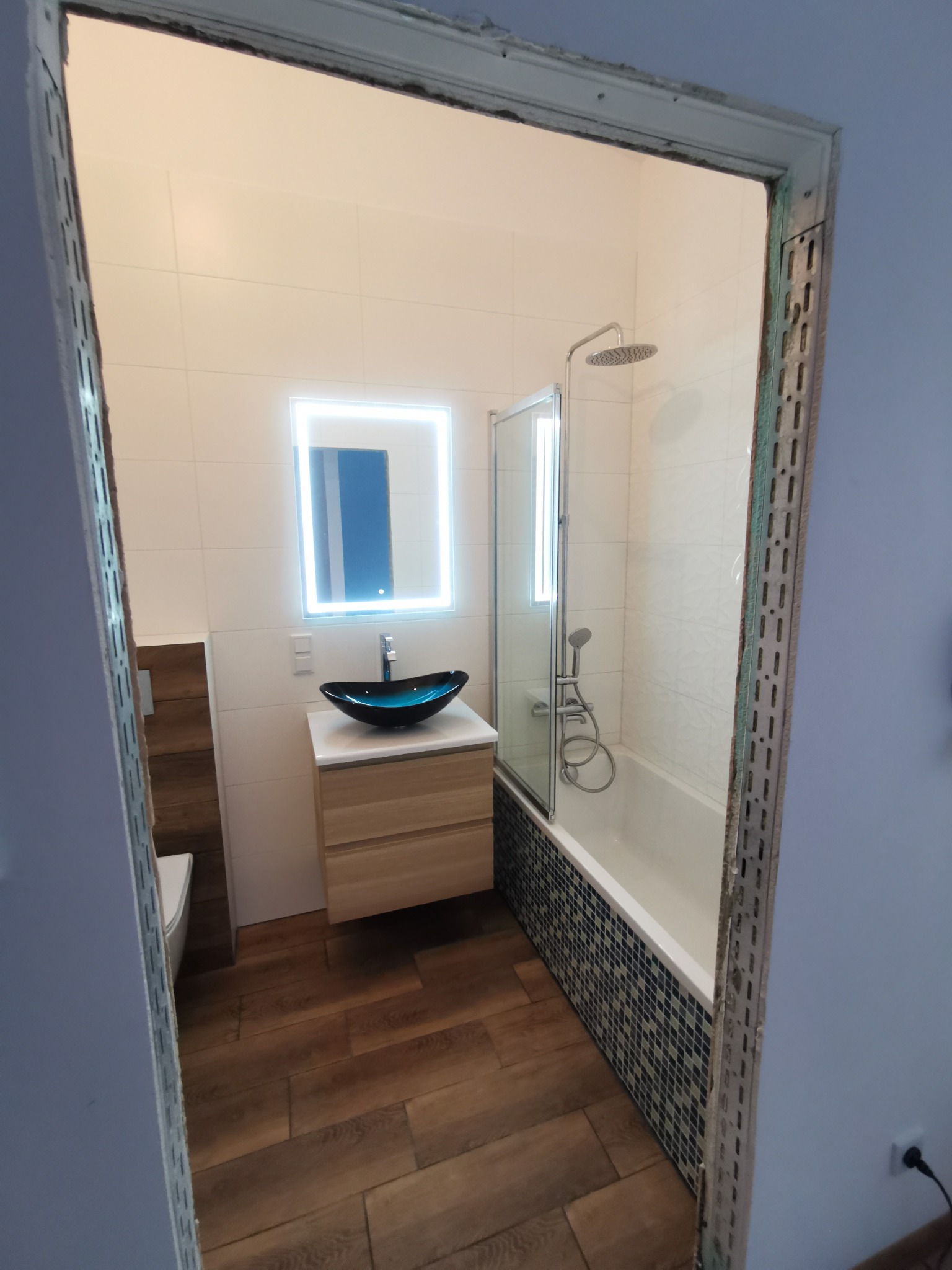 Widok na wykończoną łazienkę z kabiną prysznicową nad wanną, umywalką w kształcie misy i podświetlanym lustrem, widziany przez surową ościeżnicę przed wykończeniem.