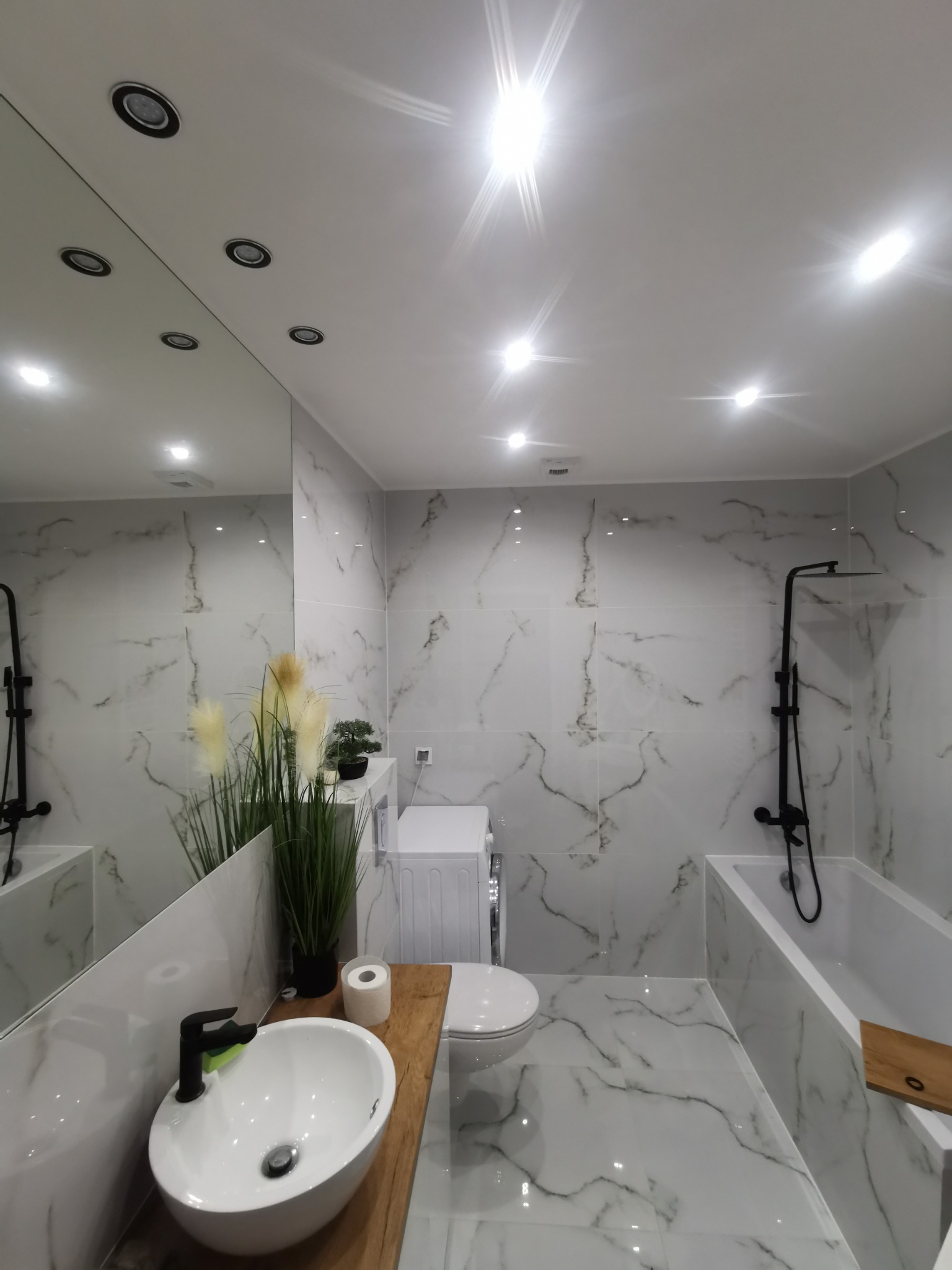 Nowoczesna łazienka z białymi płytkami imitującymi marmur, czarną armaturą, drewnianym blatem pod umywalką i dekoracyjnymi roślinami.