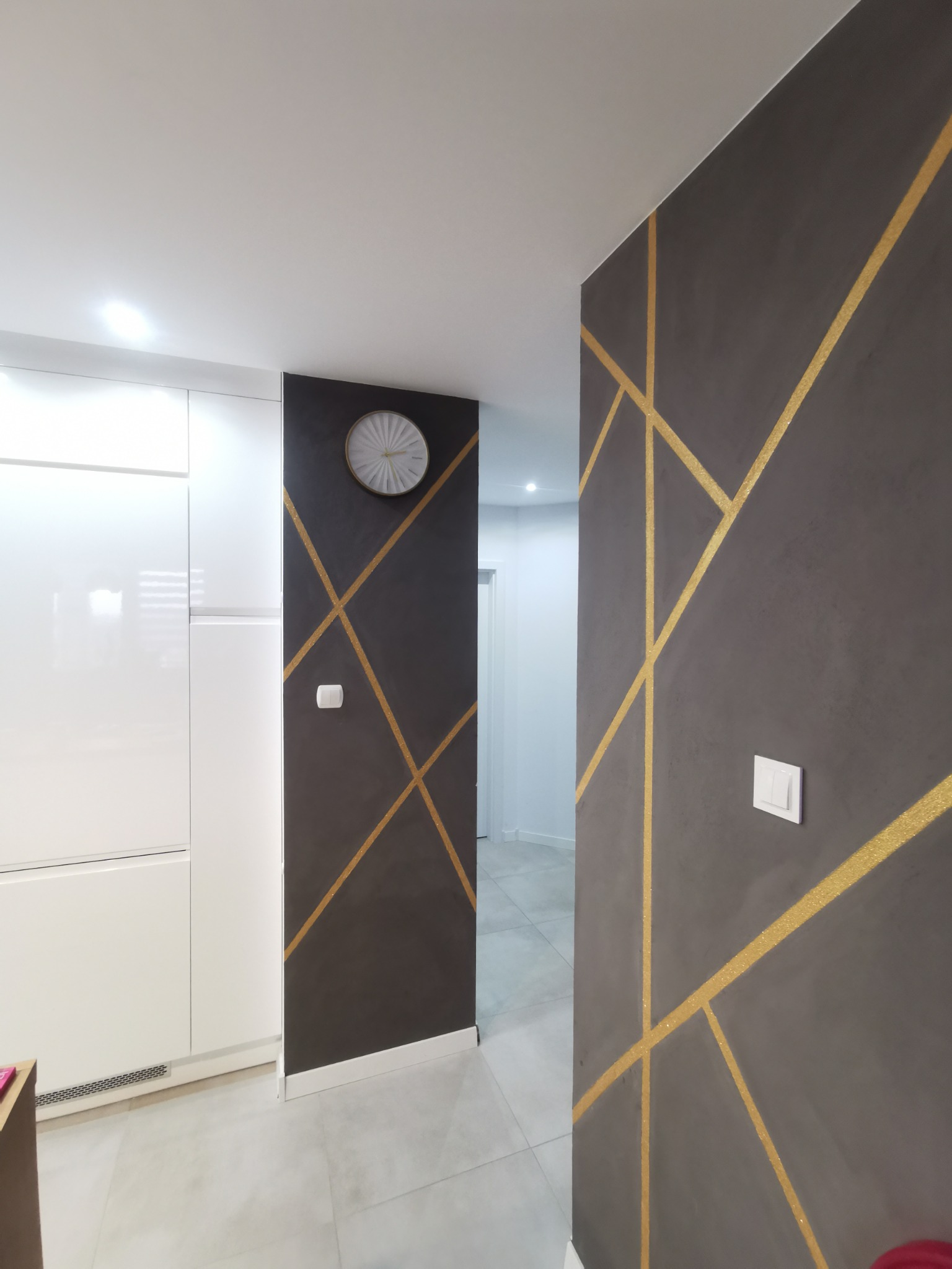 Szara ściana z geometrycznym wzorem z brokatowych, złotych linii, białe meble w tle, nowoczesny zegar na ścianie.