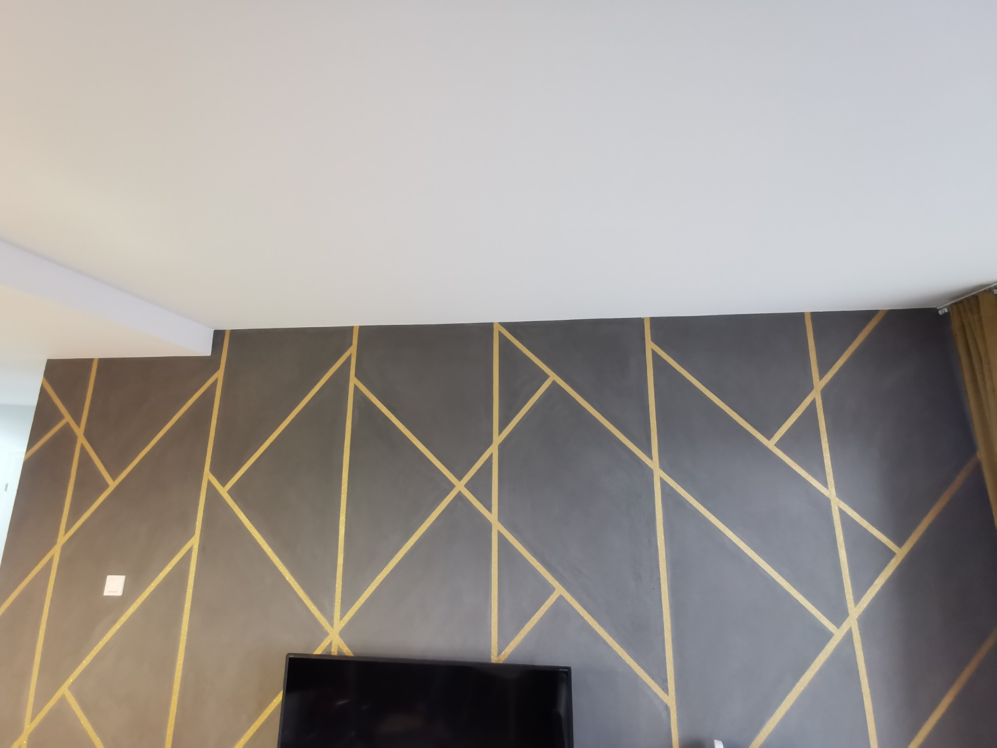 Szara ściana z geometrycznym wzorem z złotych linii w salonie, widoczny fragment sufitu i telewizora.