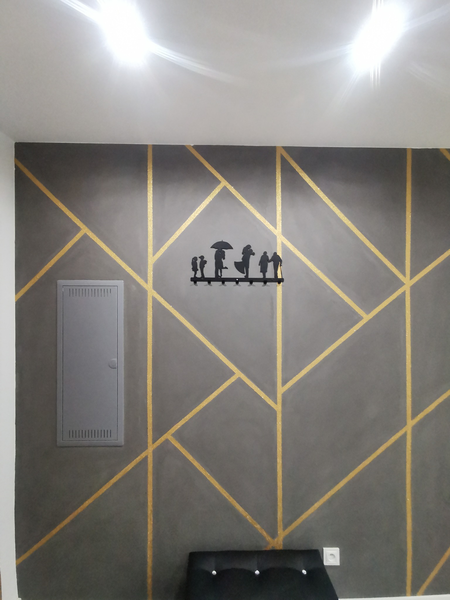 Szara ściana z geometrycznym wzorem w złotych liniach, ozdobiona metalowym wieszakiem na klucze w kształcie sylwetek ludzi pod parasolem, obok skrzynka elektryczna.