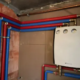 Nowoczesny piec gazowy z manometrami, podłączony do instalacji z rur w kolorach czerwonym i niebieskim, zamontowany na brązowych płytkach w kotłowni.