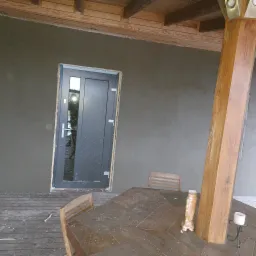 Świeżo otynkowana szara elewacja domu z drewnianą konstrukcją dachu i stolarką okienną w kolorze grafitowym, widoczne ślady zaprawy na stole i krzesłach.