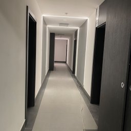 Usługi remontowo budowlane Jędrocha - Długi, jasny korytarz w nowoczesnym stylu z białymi ścianami, szarą podłogą, czarnymi futrynami drzwi i liniowym oświetleniem LED wzdłuż ścian.