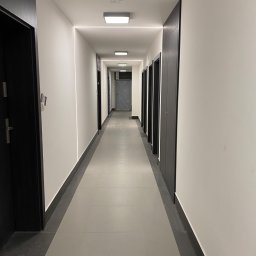 Usługi remontowo budowlane Jędrocha - Długi korytarz w nowoczesnym stylu z rzędem ciemnych drzwi po obu stronach, oświetlony liniami LED w suficie i listwami przypodłogowymi w kolorze grafitowym.