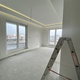 Usługi remontowo budowlane Jędrocha - Wnętrze w trakcie prac wykończeniowych: surowe ściany, betonowa podłoga, aluminiowa drabina oparta o ścianę, okna balkonowe z widokiem na miasto, podwieszany sufit z oświetleniem LED.