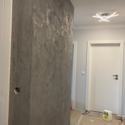 Usługi remontowo budowlane Jędrocha - Korytarz w trakcie prac wykończeniowych z dekoracyjną ścianą o szarej, nieregularnej fakturze, widoczne białe drzwi z czarną klamką i puszką elektryczną w ścianie. Na podłodze zabezpieczenia...