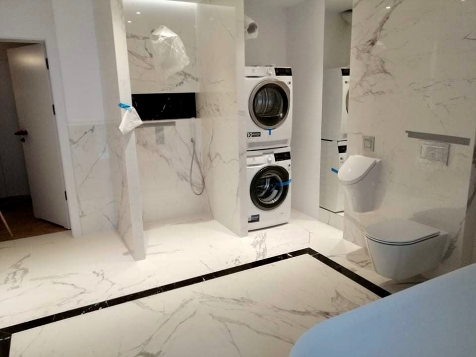 Nowoczesna łazienka z marmurowymi płytkami, wiszącą toaletą i pralko-suszarką w zabudowie, widok na prysznic z czarnym panelem.