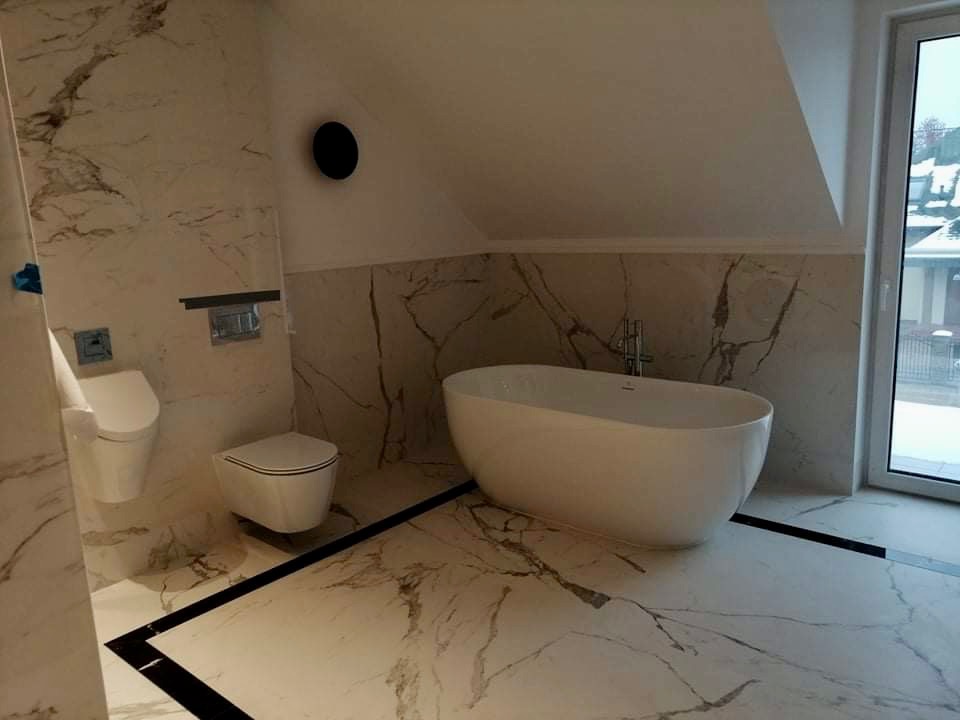 Elegancka łazienka na poddaszu z marmurowymi płytkami, wolnostojąca biała wanna i podwieszana toaleta z czarnymi akcentami.