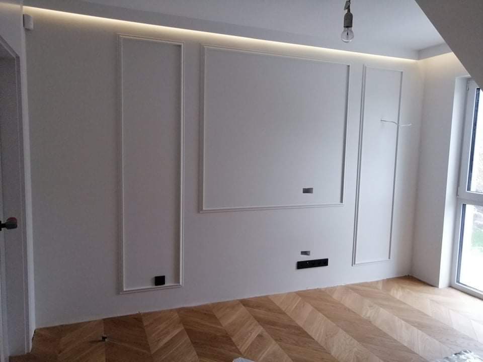 Białe ściany z dekoracyjnymi listwami i podświetleniem LED na suficie, parkiet ułożony w jodełkę, widoczny fragment okna i drzwi.