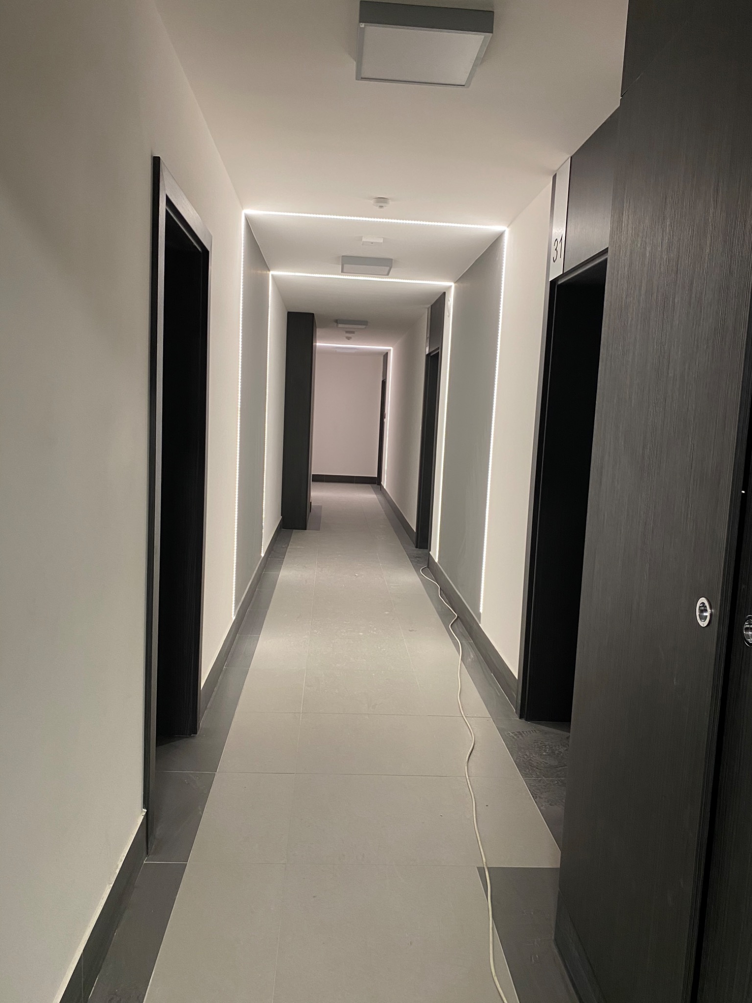 Długi, jasny korytarz w nowoczesnym stylu z białymi ścianami, szarą podłogą, czarnymi futrynami drzwi i liniowym oświetleniem LED wzdłuż ścian.