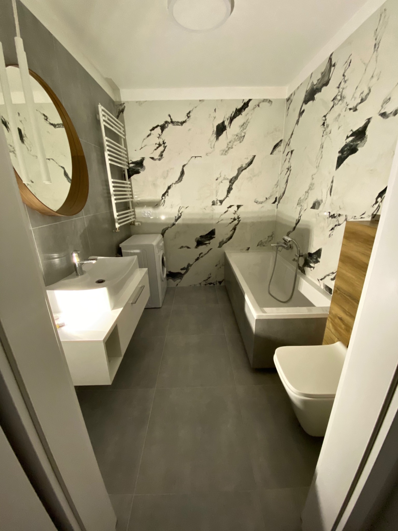 Nowoczesna łazienka po remoncie z marmurowymi płytkami na ścianach, umywalką z szafką, grzejnikiem drabinkowym, wanną i toaletą.