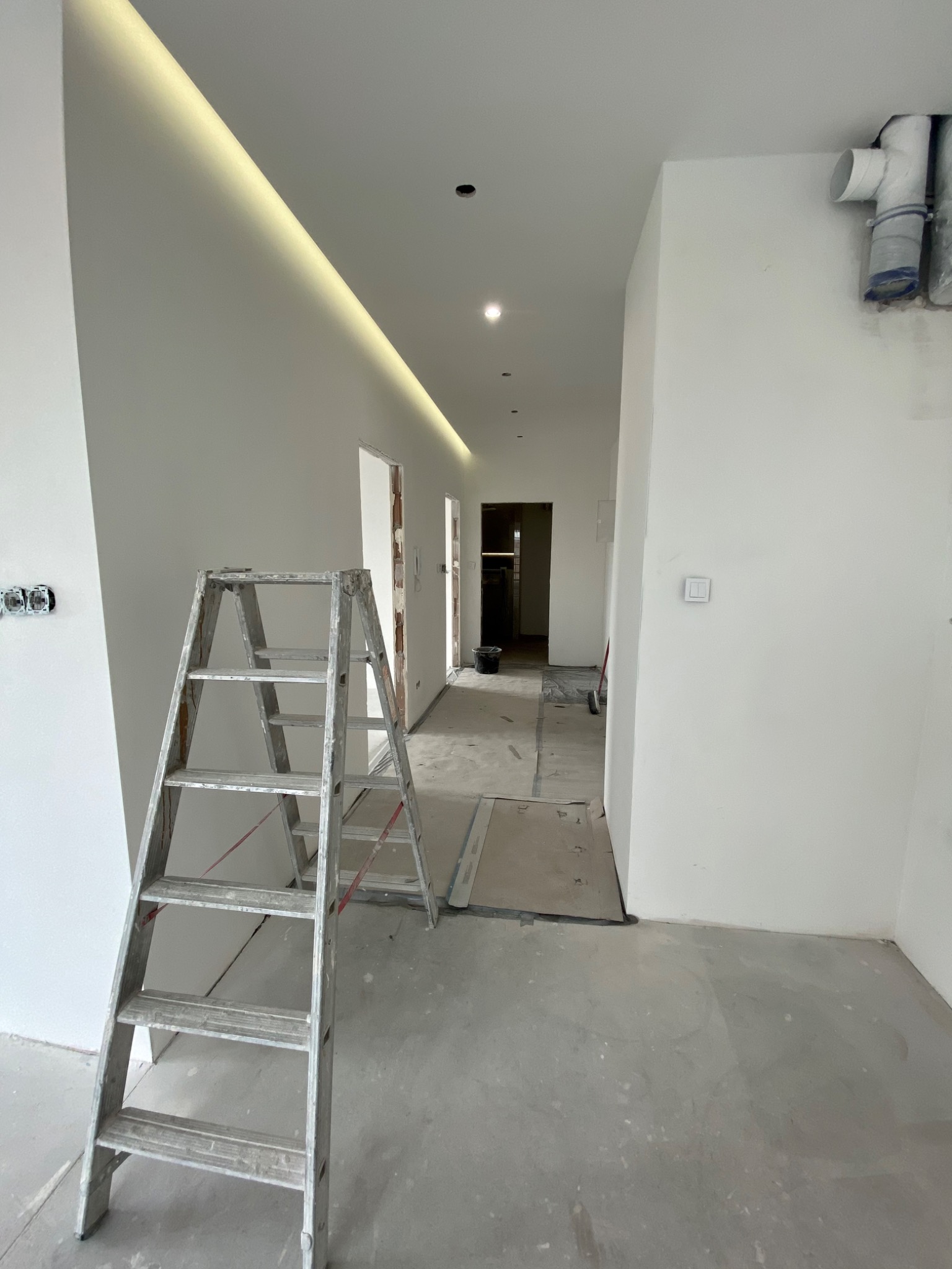 Korytarz w trakcie prac wykończeniowych, widoczna drabina aluminiowa, gładkie białe ściany z oświetleniem LED w suficie, odkryte instalacje elektryczne i rury wentylacyjne.