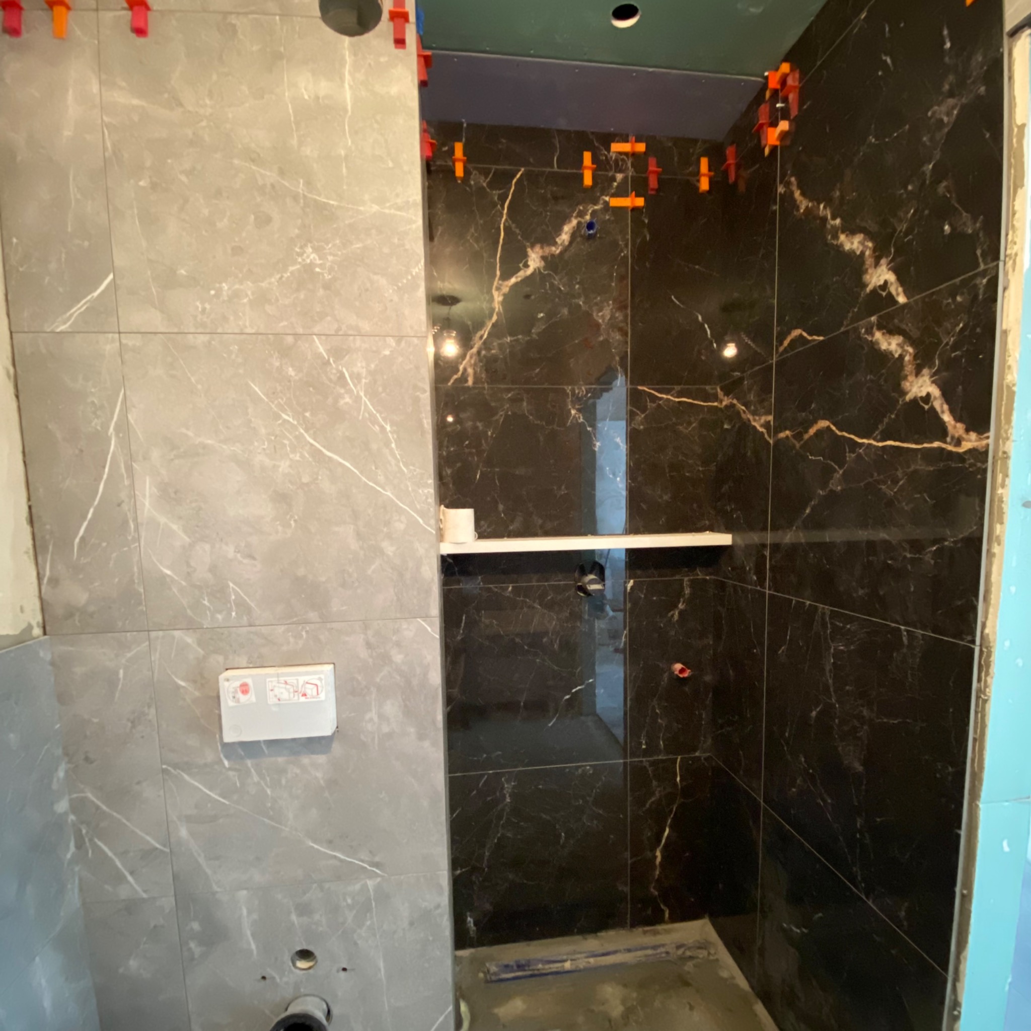 Częściowo wykończona łazienka z szarymi i czarnymi płytkami imitującymi marmur, z widocznymi klipsami poziomującymi płytki i półką w kabinie prysznicowej.