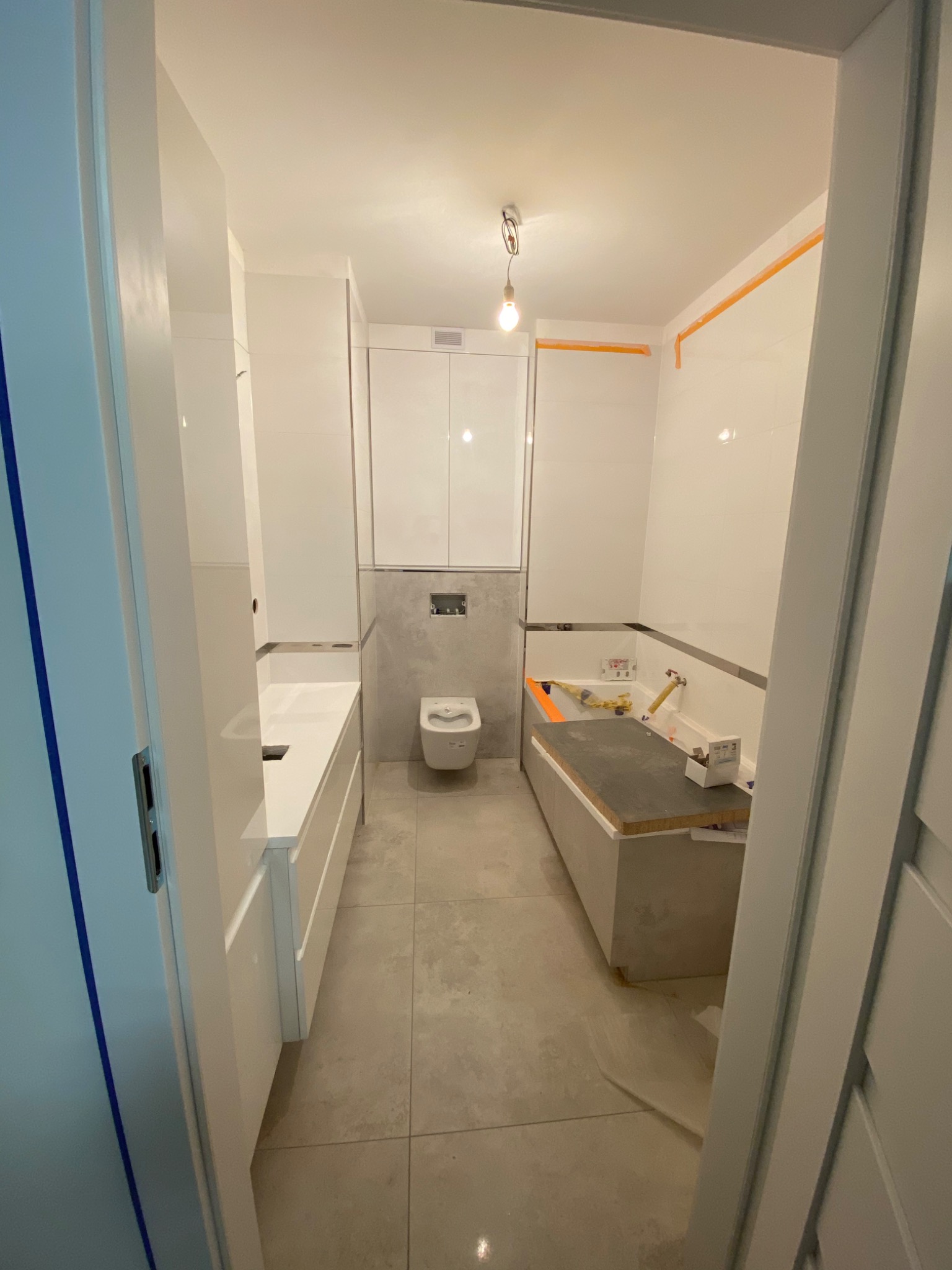 Wnętrze łazienki w trakcie remontu, widoczna zabudowa wanny z szarą płytą, białe płytki na ścianach z pomarańczową taśmą zabezpieczającą, biała umywalka z szafkami, wisząca toaleta, oraz narzędzia...