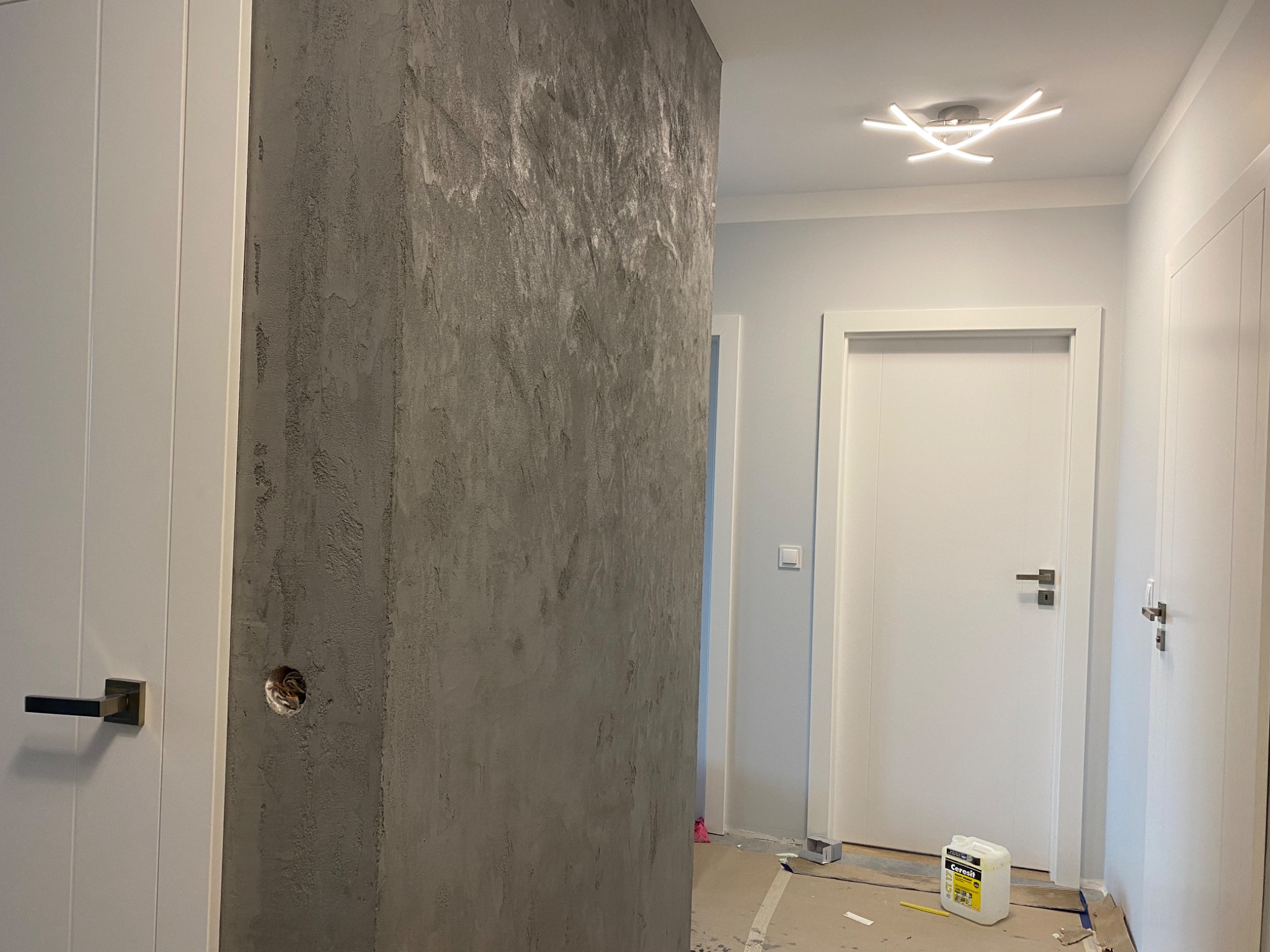 Korytarz w trakcie prac wykończeniowych z dekoracyjną ścianą o szarej, nieregularnej fakturze, widoczne białe drzwi z czarną klamką i puszką elektryczną w ścianie. Na podłodze zabezpieczenia...