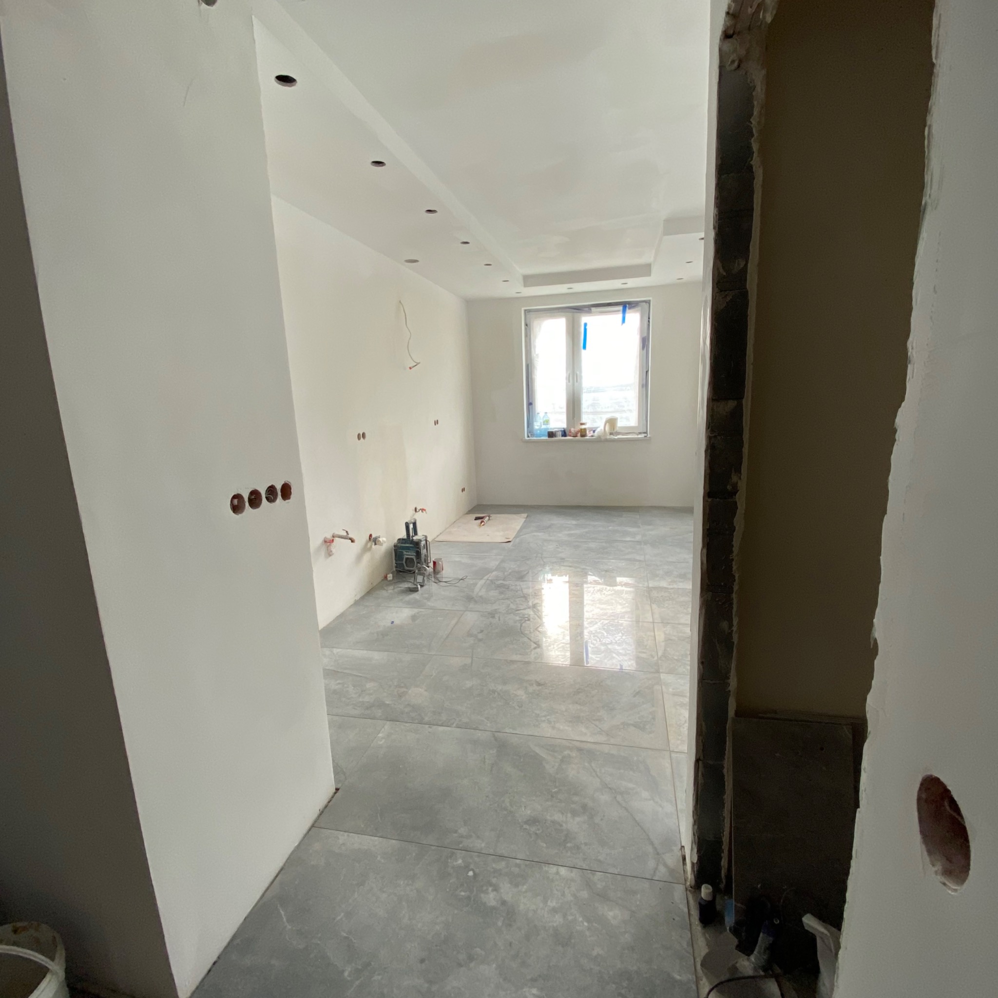 Wnętrze w trakcie remontu, widoczne szare płytki na podłodze, ściany przygotowane pod malowanie z widocznymi otworami na gniazdka elektryczne, okno z zabezpieczeniami, sufit z oświetleniem...