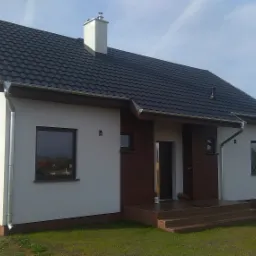 Gotowy dom szkieletowy z ciemną dachówką, białymi ścianami i drewnianą elewacją, obok widać budowę z czerwonej cegły.