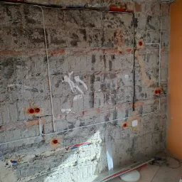 Surowa ściana z cegły i betonu w trakcie instalacji elektrycznej, widoczne kable w peszlach, puszki instalacyjne, oraz rury na podłodze w nieukończonym pomieszczeniu.