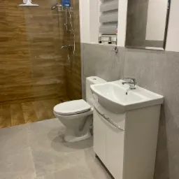 Nowoczesna łazienka z kabiną prysznicową z brązowymi płytkami imitującymi drewno, białą umywalką z szafką i toaletą, szare płytki na ścianach i podłodze, oraz akcesoria łazienkowe.