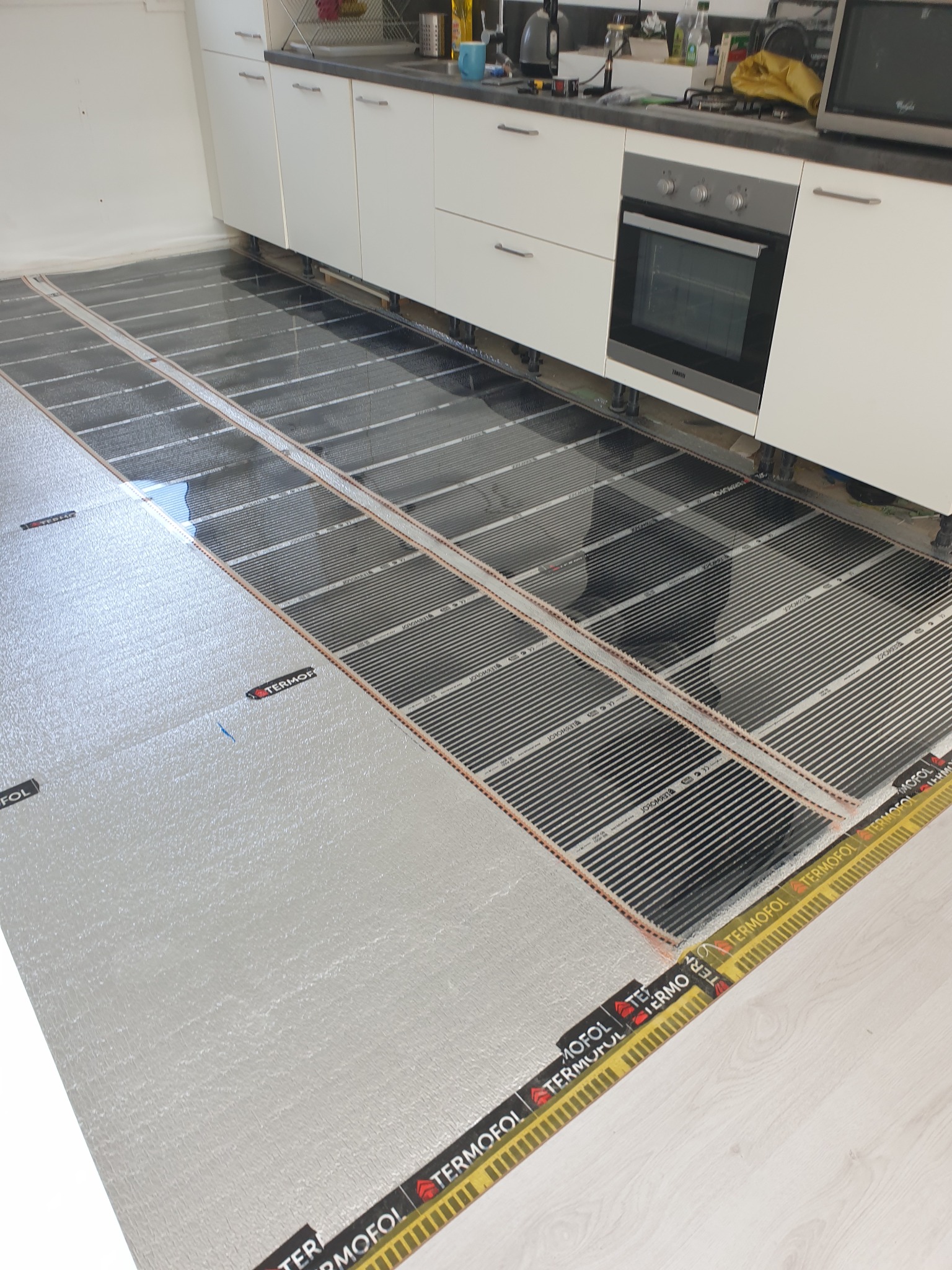Montaż folii grzewczej Termofol na podłodze w kuchni, widoczne białe szafki kuchenne i piekarnik w tle, częściowo położona srebrna folia izolacyjna.