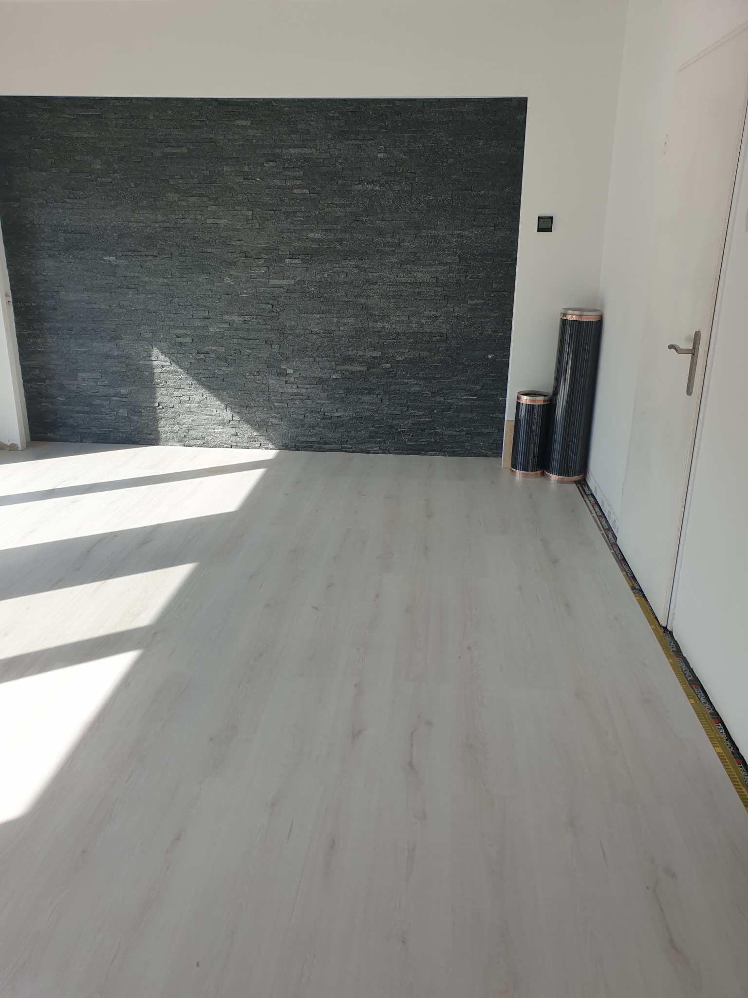Jasne panele podłogowe w nowym pomieszczeniu z fragmentem ściany wykończonej czarnym kamieniem dekoracyjnym i zwiniętą folią grzewczą przy ścianie.