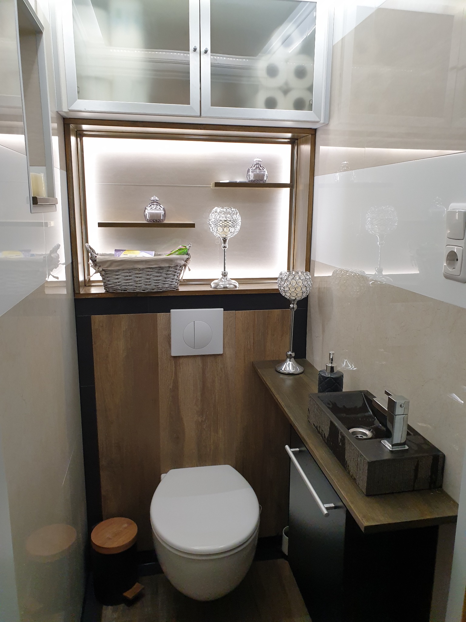 Nowoczesna aranżacja małej łazienki z podwieszaną toaletą, umywalką nablatową o fakturze kamienia, drewnopodobną zabudową i dekoracyjnymi elementami oświetleniowymi.