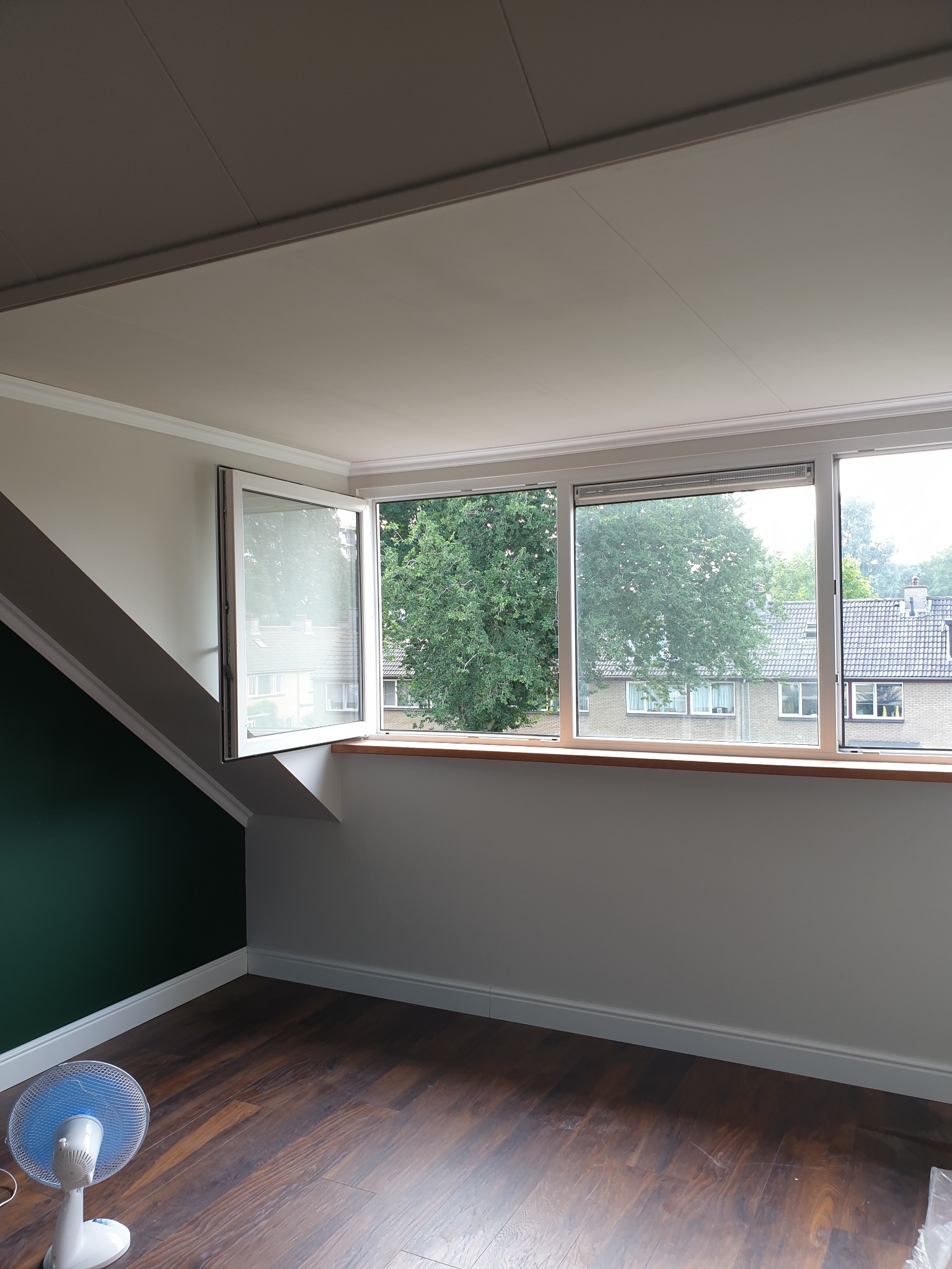 Pokój na poddaszu z drewnianą podłogą, otwartym oknem z widokiem na zieleń i fragmentem ściany pomalowanej na ciemnozielono, z wentylatorem stojącym w rogu.