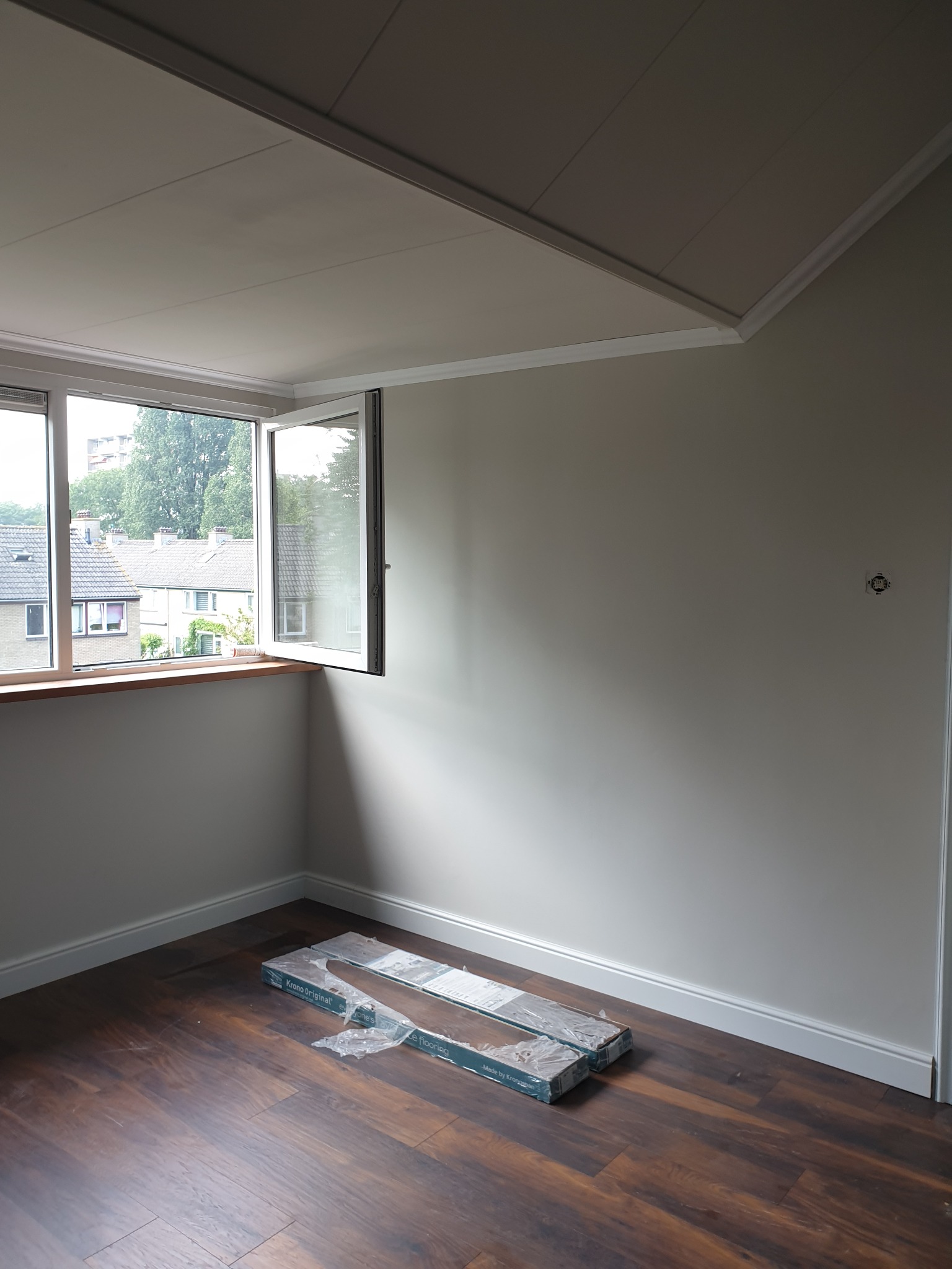 Pokój z brązową podłogą, jasnymi ścianami i otwartym oknem, na podłodze leżą paczki paneli podłogowych, przygotowane do ułożenia.