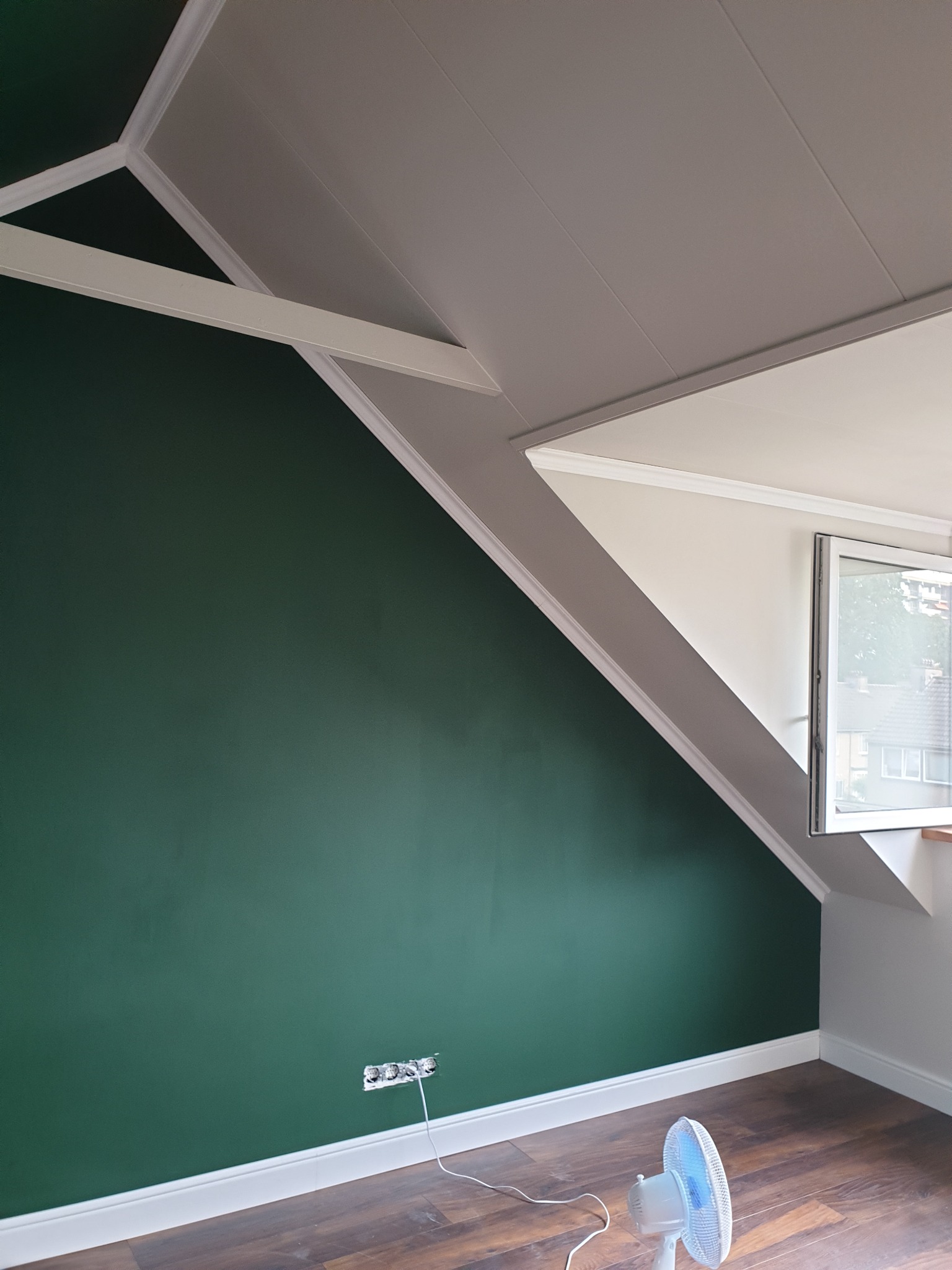 Pomieszczenie na poddaszu z zieloną ścianą, brązowymi panelami podłogowymi i białymi listwami przypodłogowymi. Widoczne gniazdka elektryczne bez osprzętu i mały wentylator podłogowy.