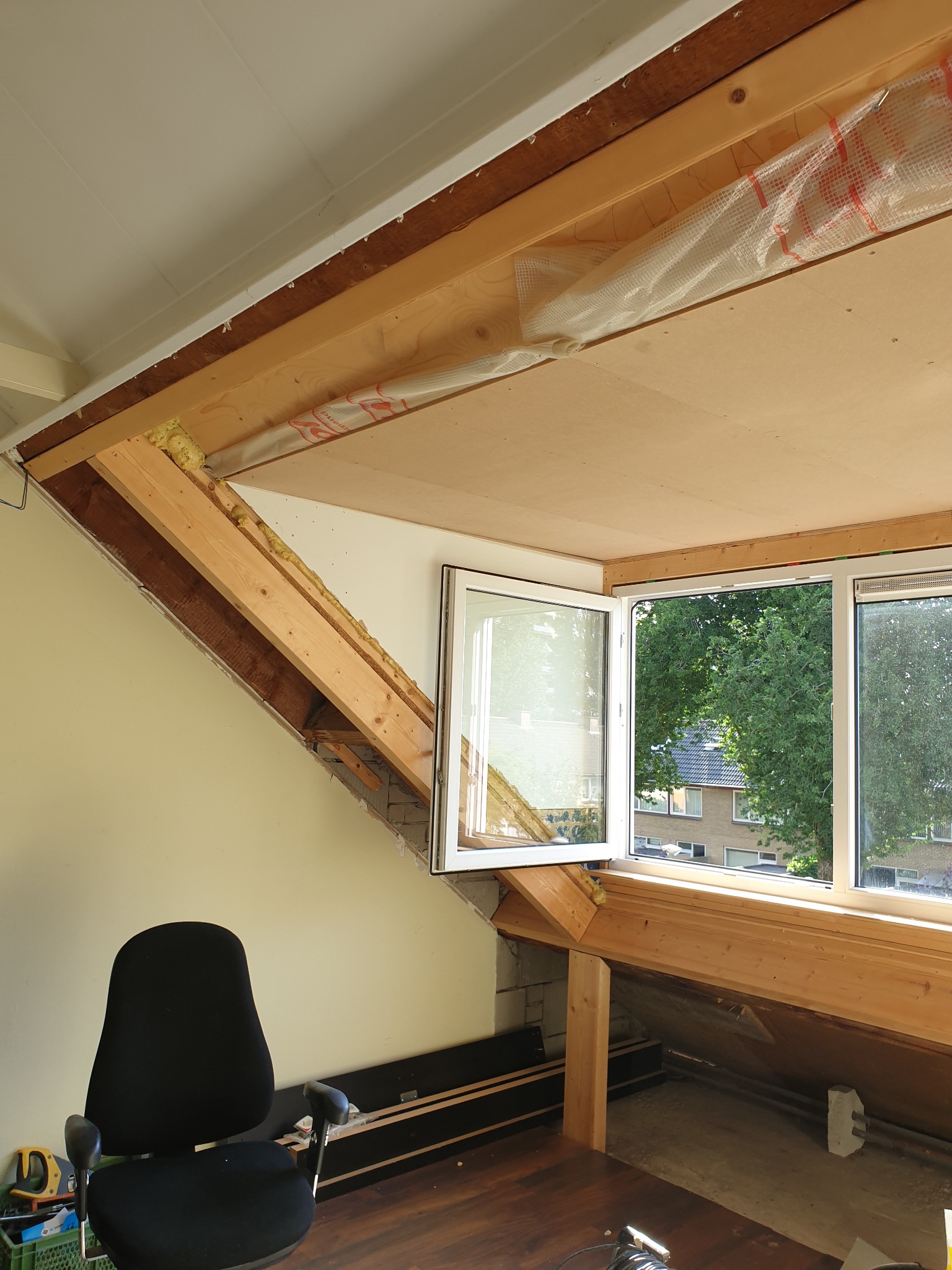 Adaptacja poddasza: widok na drewnianą konstrukcję dachu z oknami, częściowo ocieploną, z widoczną wełną mineralną i folią ochronną, z przygotowaną podłogą z ciemnego drewna oraz biurkiem...