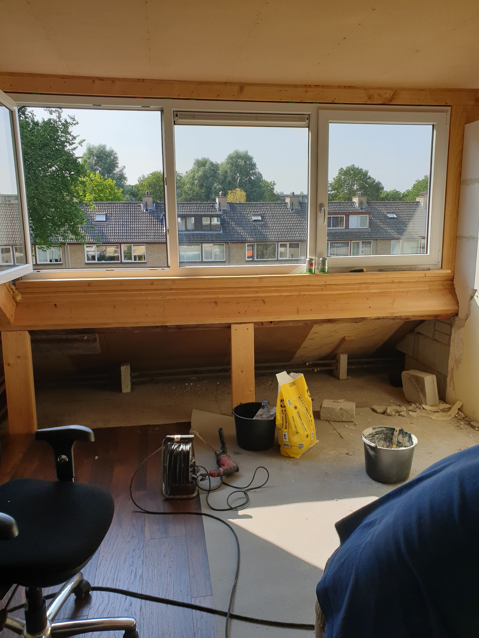 Adaptacja poddasza: widok na okna dachowe z drewnianą konstrukcją pod parapet, narzędzia pracy i materiały budowlane w nieukończonym pomieszczeniu.