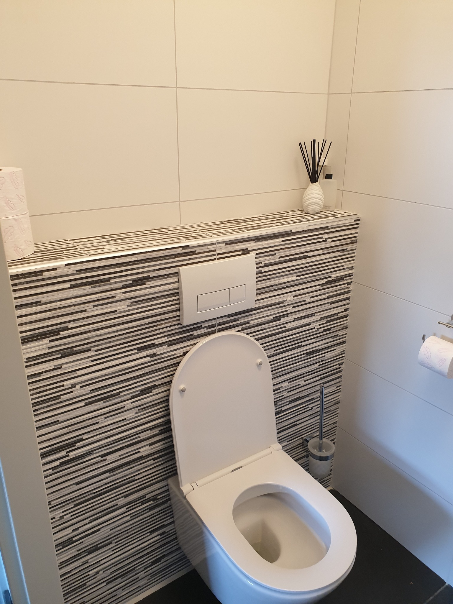 Nowoczesna toaleta z podwieszanym sedesem, mozaiką w odcieniach szarości i białymi płytkami, uzupełniona o zapas papieru toaletowego i odświeżacz powietrza w dekoracyjnym wazonie.