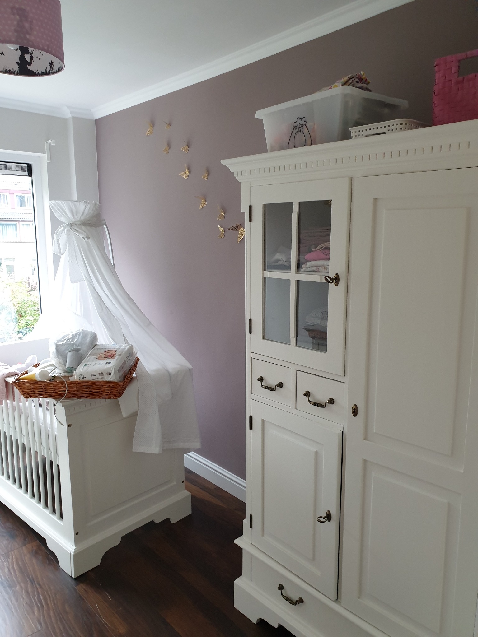 Pokój dziecięcy z białym łóżeczkiem z baldachimem, wiklinowym koszem z akcesoriami dla niemowląt i dużą białą szafą z przeszklonymi drzwiczkami na tle liliowej ściany zdobionej złotymi motylami.