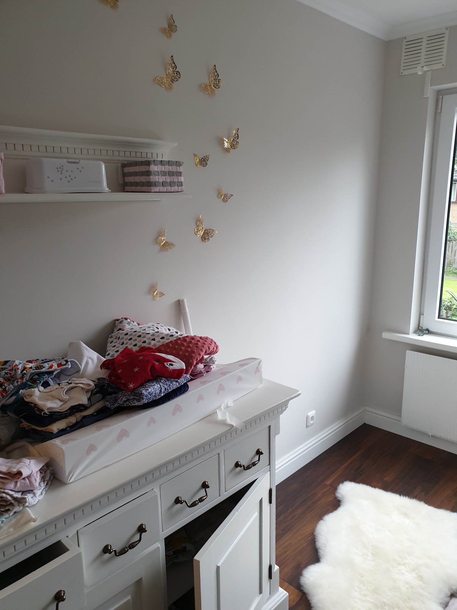Pokój dziecięcy po remoncie: biała komoda z przewijakiem, na niej sterta ubranek, na ścianie złote motyle, drewniana podłoga z białym dywanikiem, półka z dekoracjami.
