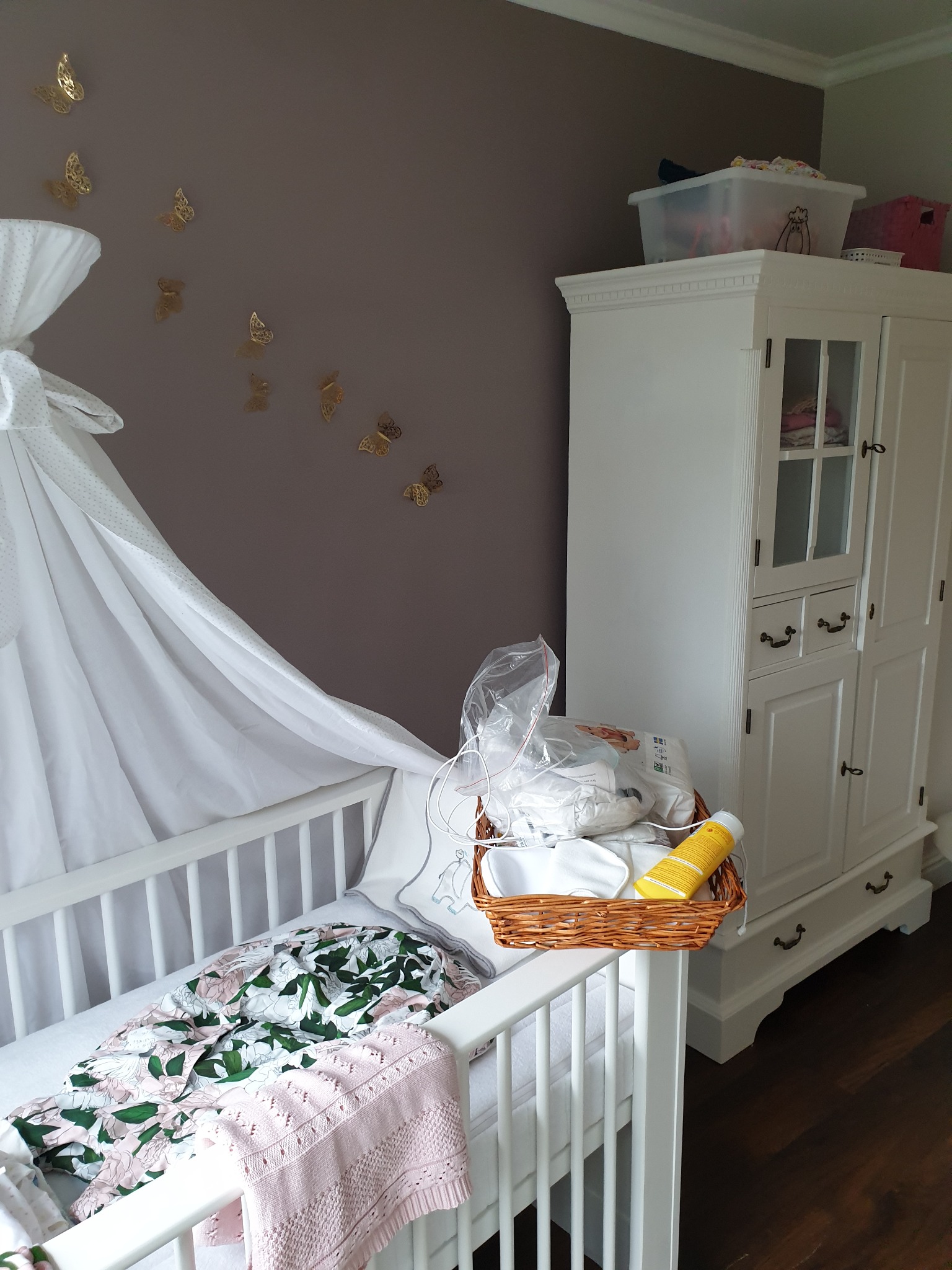 Pokój dziecięcy z białym łóżeczkiem z baldachimem, na którym leży kocyk w kwiaty i różowa narzuta, obok biała szafa i ściana ozdobiona złotymi motylami.