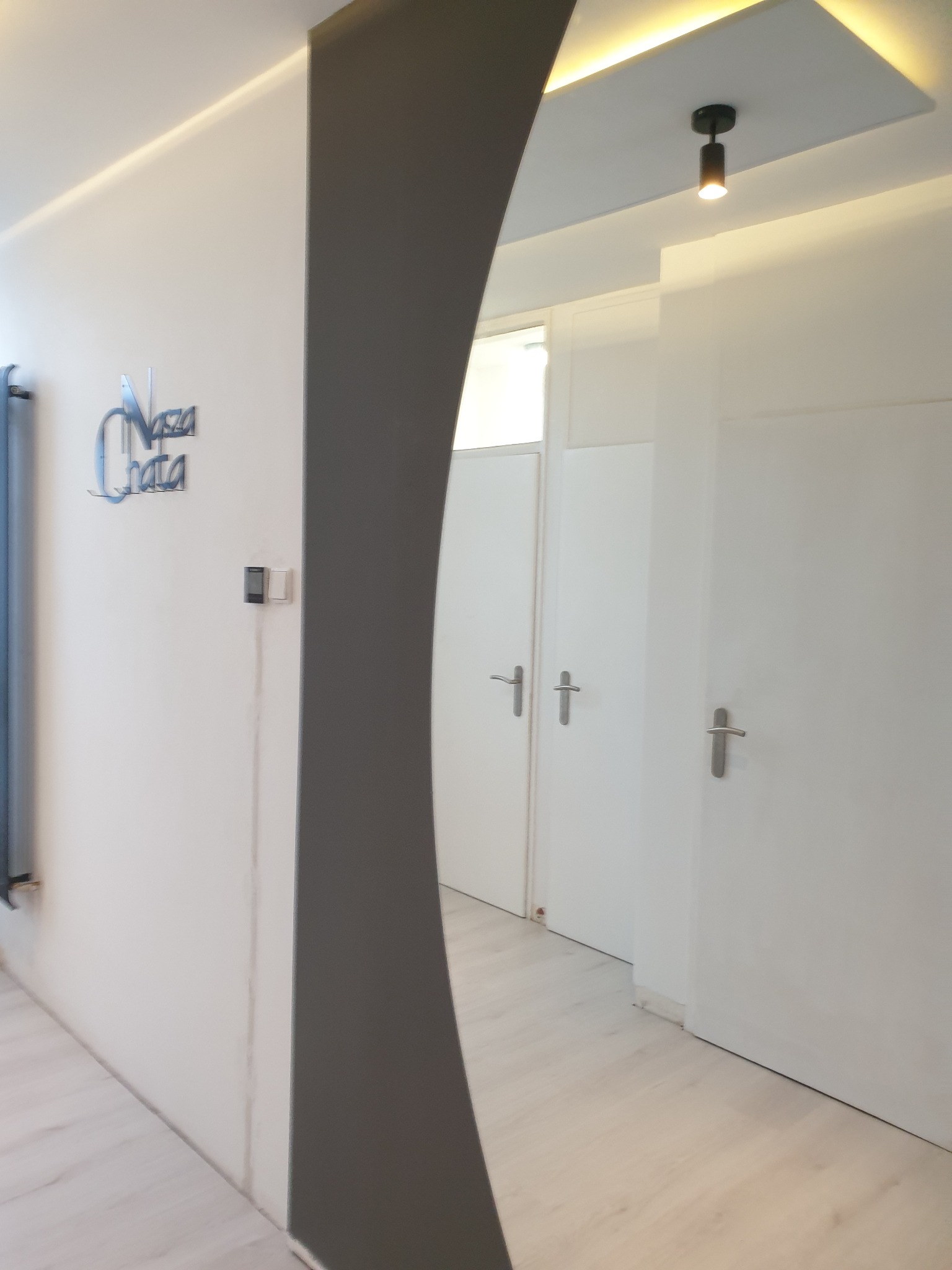 Nowoczesne wnętrze przedpokoju z białymi ścianami, jasną podłogą, szarą dekoracyjną kolumną i podświetlanym sufitem, widoczne drzwi wewnętrzne z srebrnymi klamkami oraz logo firmy na ścianie.