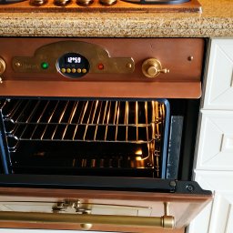 Serwis AGD w Szczecine - Otwarty piekarnik w stylu retro z miedzianym wykończeniem, widoczne wnętrze z rusztem, białe szafki kuchenne z ozdobnymi uchwytami w tle.