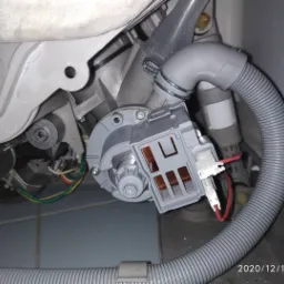 Wnętrze zmywarki z widoczną pompą obiegową, wężem odpływowym i elementami elektrycznymi, ukazujące proces naprawy lub wymiany części.