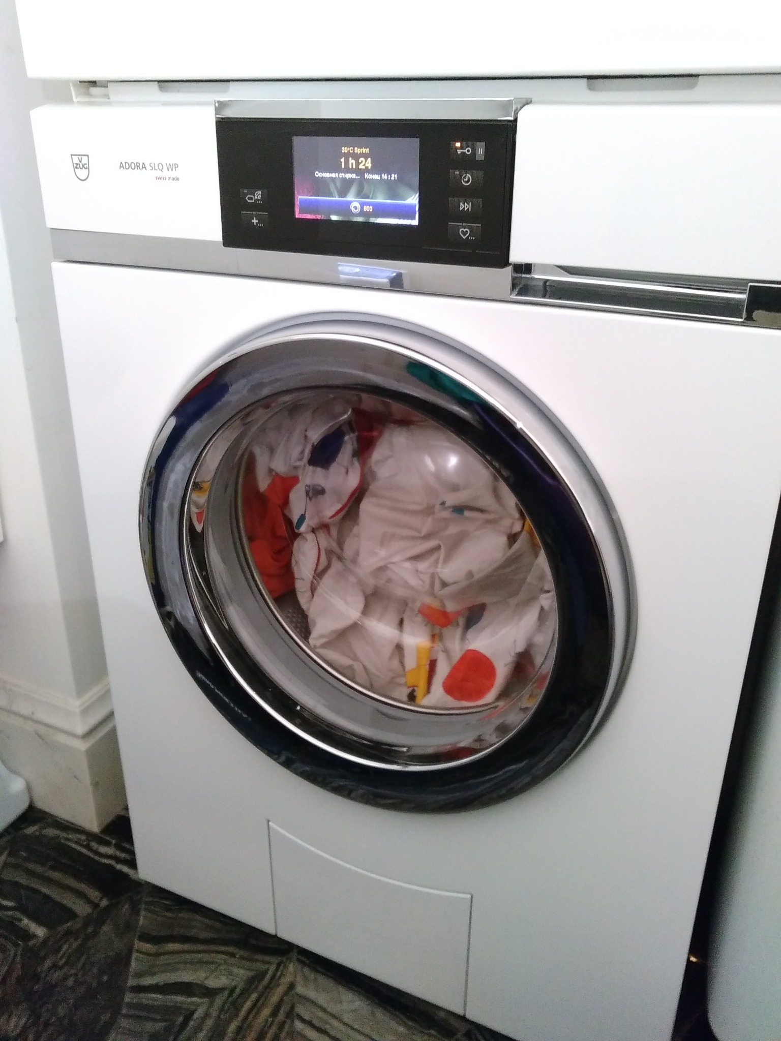 Pralka automatyczna marki Adora SLQ WP z załadunkiem od przodu, wypełniona częściowo kolorowym praniem, widoczny panel sterowania z wyświetlaczem wskazującym czas trwania programu.