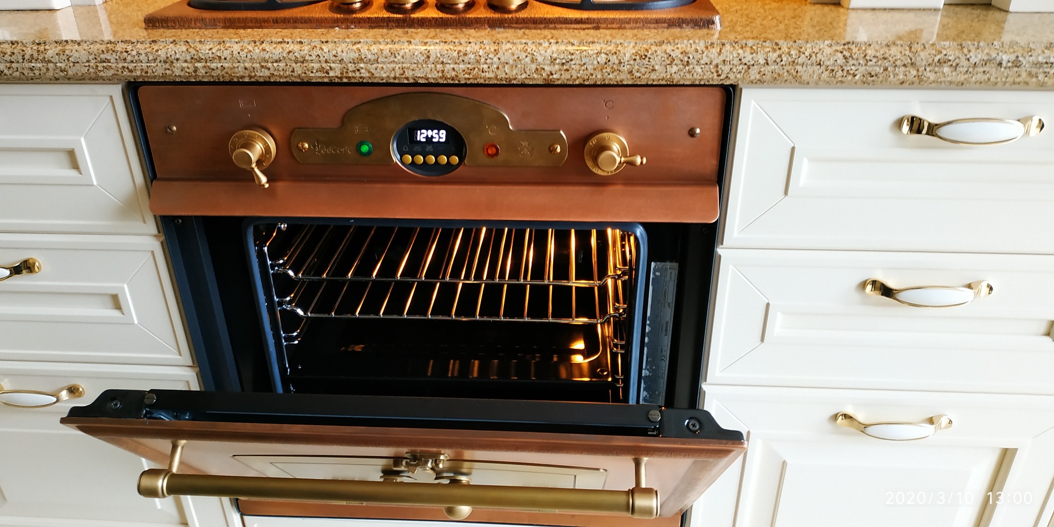 Otwarty piekarnik w stylu retro z miedzianym wykończeniem, widoczne wnętrze z rusztem, białe szafki kuchenne z ozdobnymi uchwytami w tle.