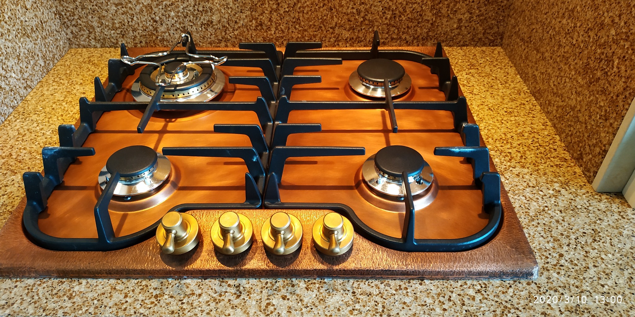 Miedziana płyta gazowa z czarnymi rusztami i mosiężnymi pokrętłami na blacie w kuchni.