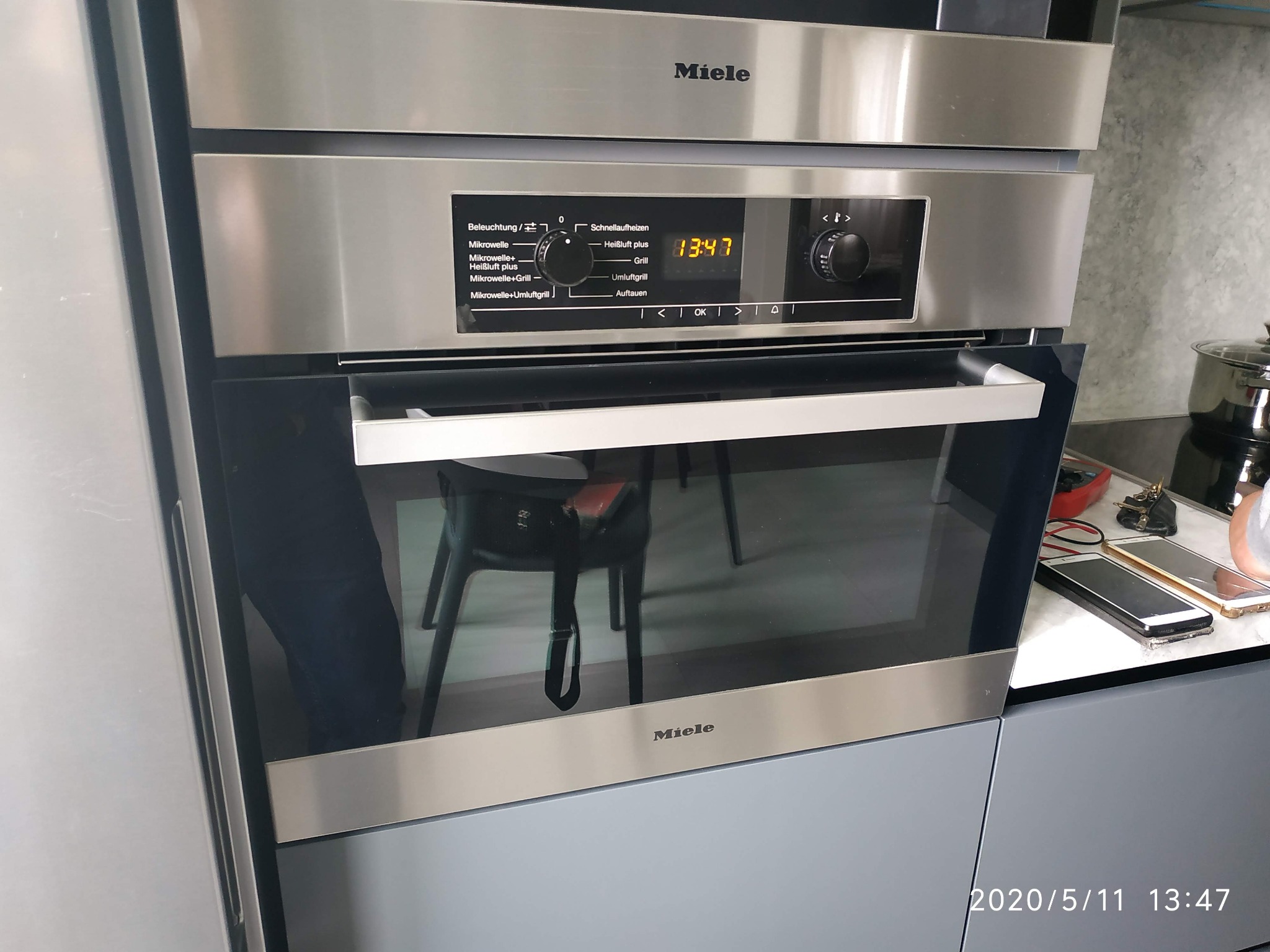 Wbudowany piekarnik Miele ze stali nierdzewnej z wyświetlaczem pokazującym godzinę 13:47 i różnymi opcjami pieczenia, widoczny w kuchni z szarą zabudową.