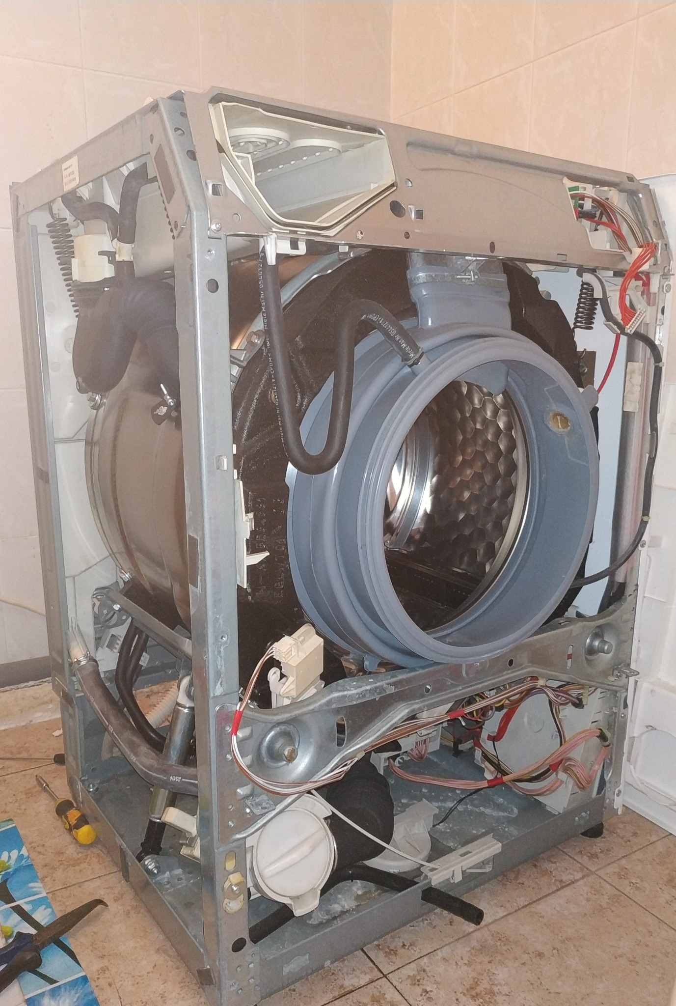 Pralka automatyczna z otwartą obudową, widoczne wnętrze bębna, elementy elektryczne i hydrauliczne, narzędzie w pobliżu