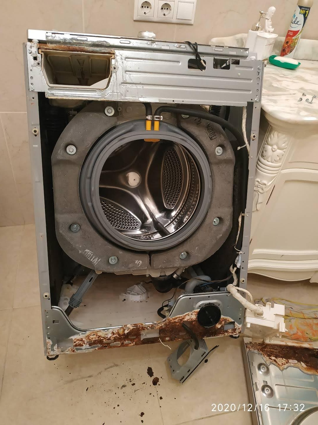 Rozłożona pralka automatyczna w trakcie naprawy, widoczne ślady korozji na obudowie, wnętrze bębna z nierdzewnej stali, elementy demontażu na podłodze w łazience.