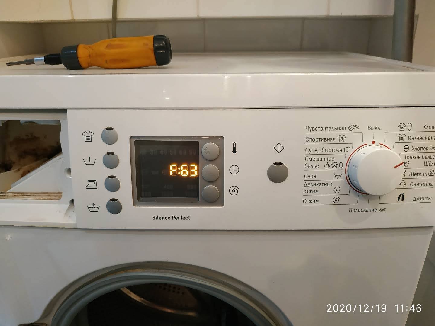 Rozkręcona pralka automatyczna z widocznym wyświetlaczem i żółtym śrubokrętem leżącym na obudowie, podczas naprawy.