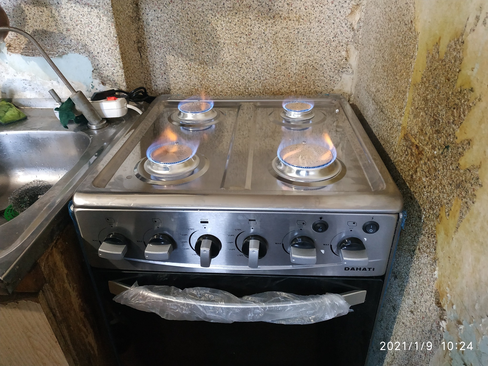 Czteropalnikowa kuchenka gazowa Dahati z zapalonymi palnikami, widoczna w bliskim planie, z fragmentem zlewu po lewej stronie i ścianą z nierówną fakturą w tle.