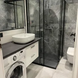 Nowoczesna łazienka z kabiną prysznicową typu walk-in z czarnymi profilami, umywalką nablatową na szafce z szufladami, pralką i płytkami imitującymi marmur.