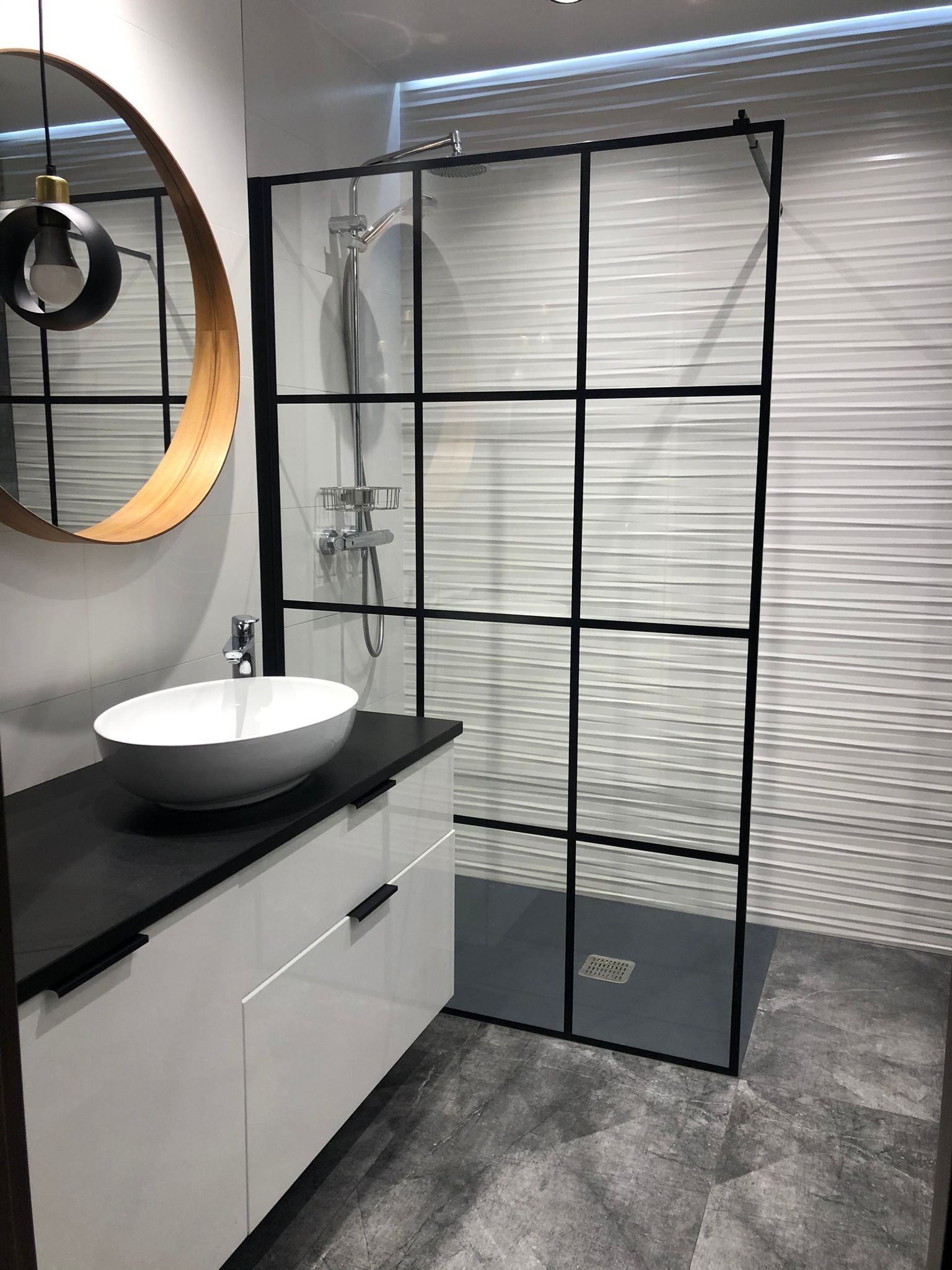 Nowoczesna łazienka z kabiną prysznicową typu walk-in z czarnymi profilami, owalną umywalką nablatową i okrągłym lustrem z drewnianą ramą.